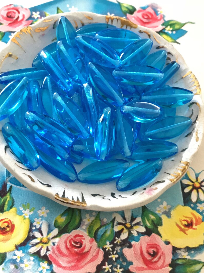 May include: A bowl filled with turquoise blue glass beads. The beads are oval shaped and have a smooth, glossy finish.