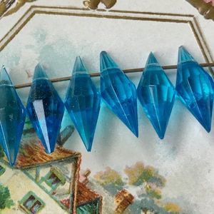 May include: A strand of turquoise blue faceted glass beads in a teardrop shape. The beads are strung on a thin wire and are arranged in a row.