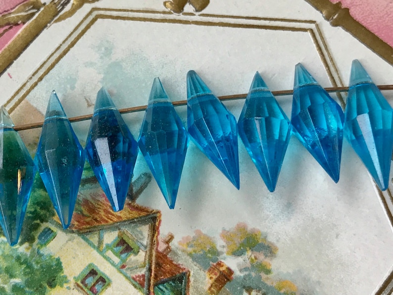 May include: A strand of turquoise blue faceted glass beads in a teardrop shape. The beads are strung on a thin wire and are arranged in a row.