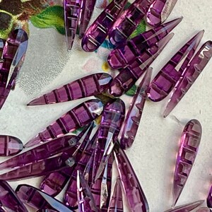 May include: A collection of purple glass teardrop-shaped beads with a faceted design. The beads are arranged in a random pattern on a white surface.