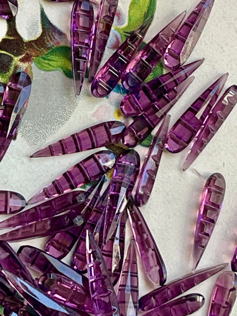 May include: A collection of purple glass teardrop-shaped beads with a faceted design. The beads are arranged in a random pattern on a white surface.