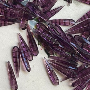 May include: A large quantity of purple glass teardrop-shaped beads. The beads have a faceted design and are arranged in a random pattern.