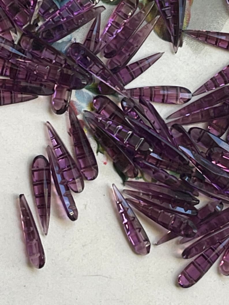 May include: A large quantity of purple glass teardrop-shaped beads. The beads have a faceted design and are arranged in a random pattern.