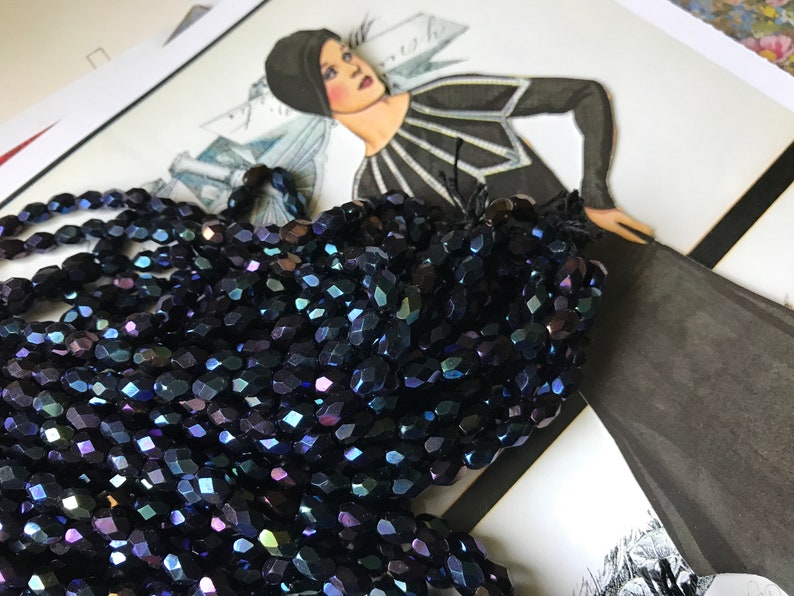May include: A strand of iridescent dark blue glass beads. The beads are faceted and have a slightly oval shape.