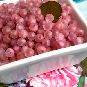May include: A bowl filled with pink frosted glass beads. The beads are round and have a slightly iridescent finish. A US penny is included for scale.