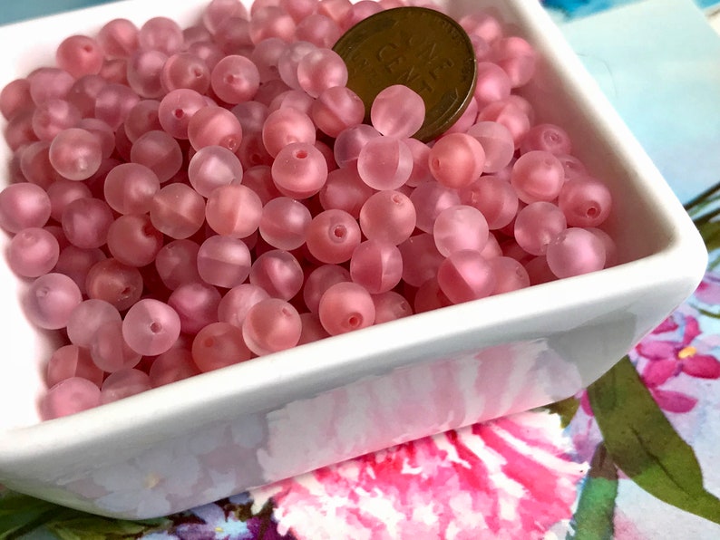 May include: A bowl filled with pink frosted glass beads. The beads are round and have a slightly iridescent finish. A US penny is included for scale.