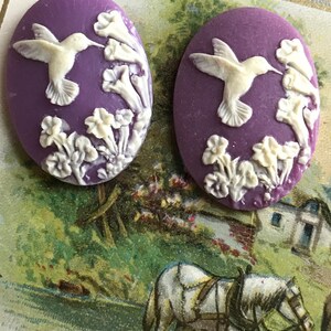 May include: Two purple cameo-style buttons with white raised designs of hummingbirds and flowers.