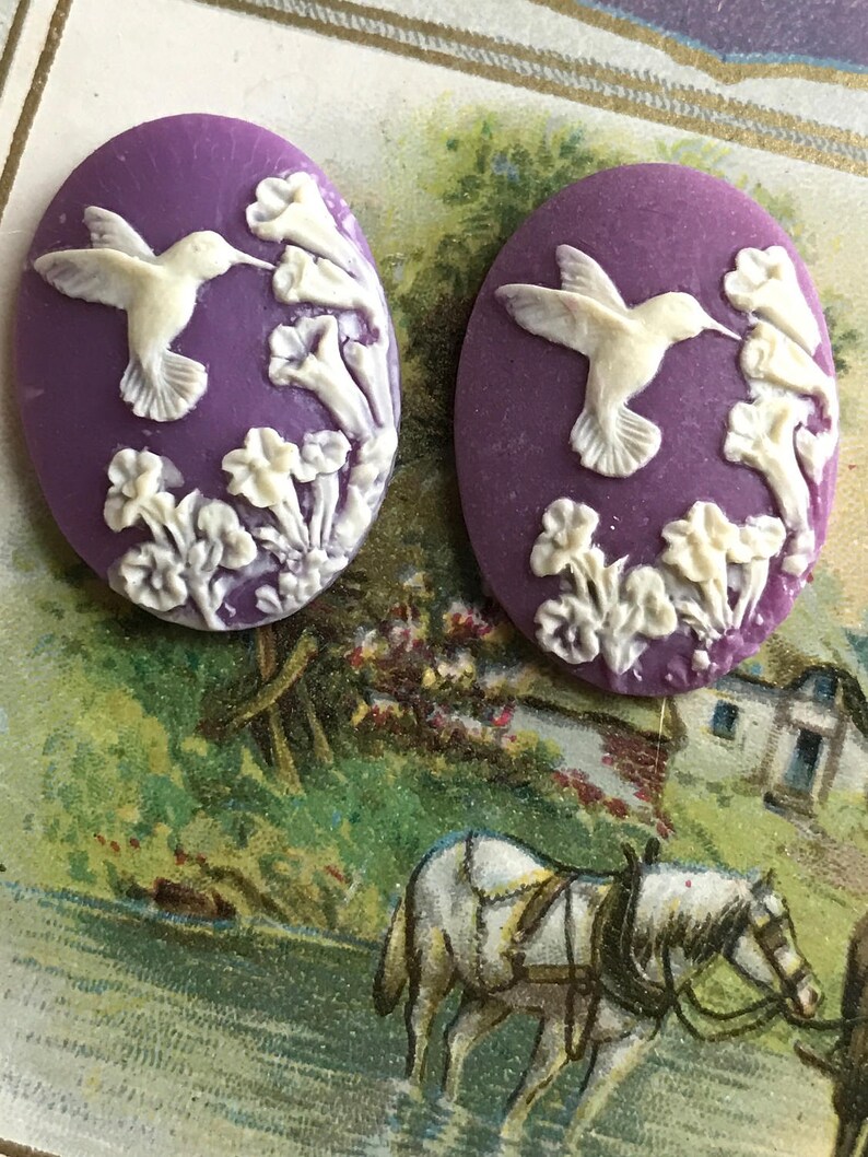 May include: Two purple cameo-style buttons with white raised designs of hummingbirds and flowers.