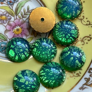 May include: A collection of eight green glass cabochons with gold flecks. The cabochons are round and have a slightly domed shape. They are set in gold-tone metal settings.