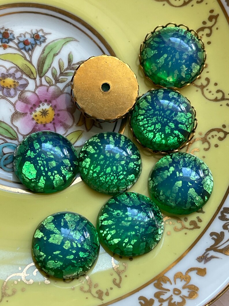 May include: A collection of eight green glass cabochons with gold flecks. The cabochons are round and have a slightly domed shape. They are set in gold-tone metal settings.