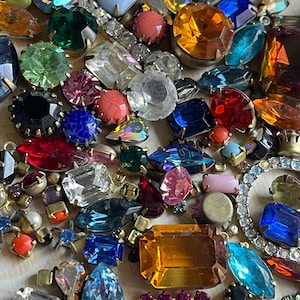 May include: A vibrant assortment of vintage rhinestones and jewels in diverse shapes and sizes. The collection features emerald green, sapphire blue, ruby red, and amber stones, with clear and iridescent accents. Some are set in gold-tone metal settings.
