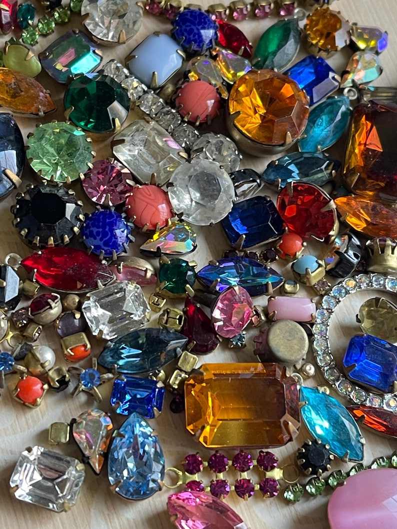 May include: A vibrant assortment of vintage rhinestones and jewels in diverse shapes and sizes. The collection features emerald green, sapphire blue, ruby red, and amber stones, with clear and iridescent accents. Some are set in gold-tone metal settings.