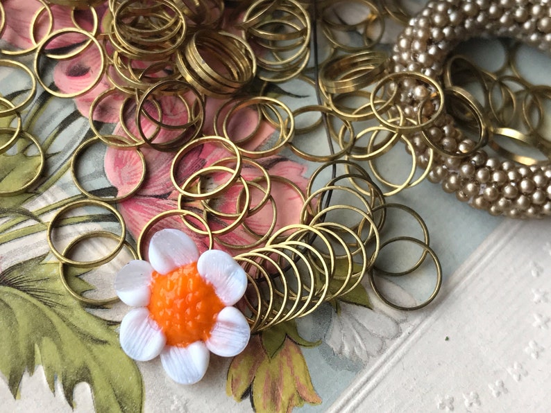 Peut inclure: Un tas d'anneaux de saut en m&eacute;tal dor&eacute;, certains empil&eacute;s et d'autres &eacute;parpill&eacute;s, avec un bouton de fleur blanc et orange au centre.