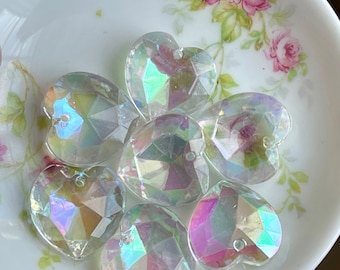 May include: A collection of iridescent clear heart-shaped glass beads. The beads have a faceted design and are arranged on a white surface.