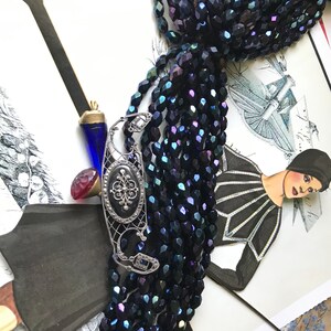 May include: A strand of dark blue iridescent glass beads with a silver filigree pendant featuring a black stone and a small diamond. The beads are arranged in a fan shape and are lying on a patterned surface.