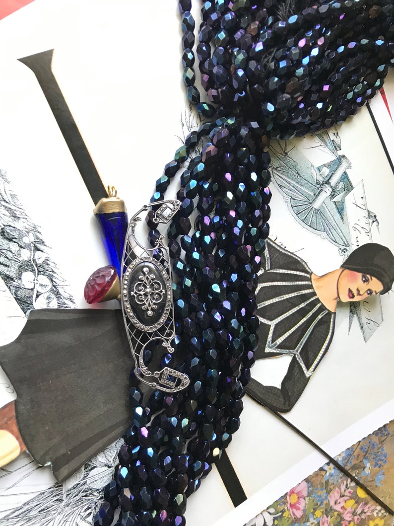May include: A strand of dark blue iridescent glass beads with a silver filigree pendant featuring a black stone and a small diamond. The beads are arranged in a fan shape and are lying on a patterned surface.