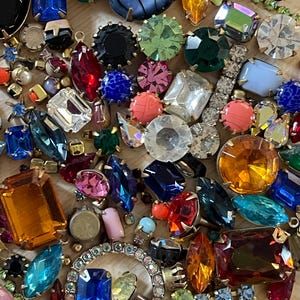 May include: A close-up of a collection of colourful gemstones in various shapes and sizes. The gems include shades of blue, green, red, orange, and clear, some set in metal. The arrangement suggests jewellery components or decorative elements.
