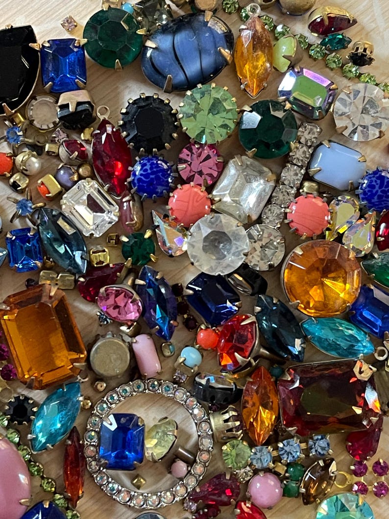 May include: A close-up of a collection of colourful gemstones in various shapes and sizes. The gems include shades of blue, green, red, orange, and clear, some set in metal. The arrangement suggests jewellery components or decorative elements.