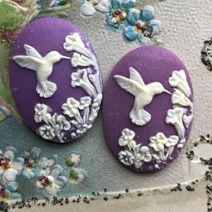 May include: Two purple oval-shaped ceramic cabochons with white raised designs of hummingbirds and flowers.