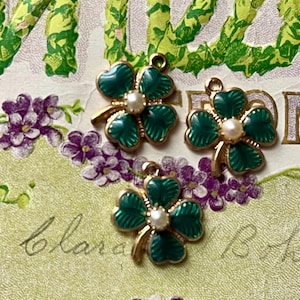 Puede incluir: Tres dijes de tr&eacute;bol verde y dorado con un centro de perla. Los dijes est&aacute;n colocados sobre un fondo vintage con flores moradas y letras verdes. Los dijes son probablemente para la fabricaci&oacute;n de joyas.