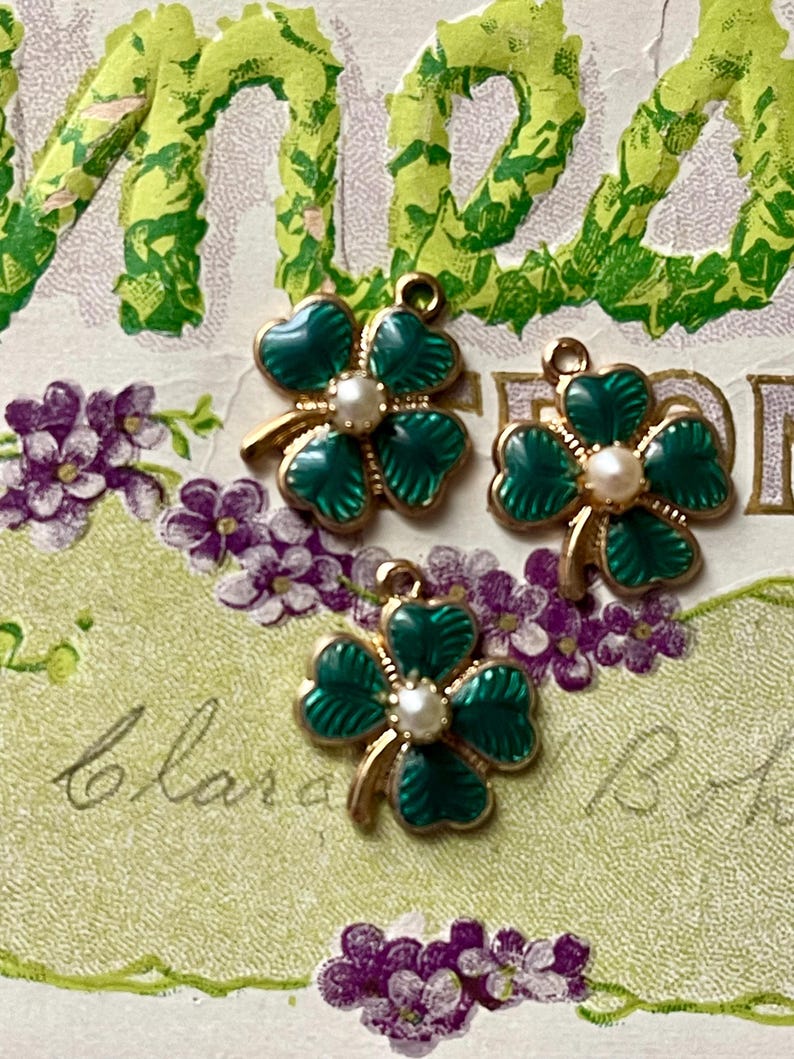 Puede incluir: Tres dijes de tr&eacute;bol verde y dorado con un centro de perla. Los dijes est&aacute;n colocados sobre un fondo vintage con flores moradas y letras verdes. Los dijes son probablemente para la fabricaci&oacute;n de joyas.