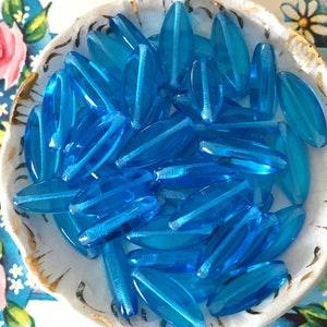 May include: A bowl filled with turquoise blue glass beads. The beads are oval shaped and have a slightly iridescent finish.