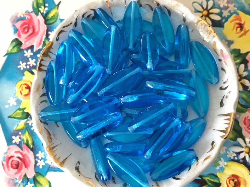 May include: A bowl filled with turquoise blue glass beads. The beads are oval shaped and have a slightly iridescent finish.