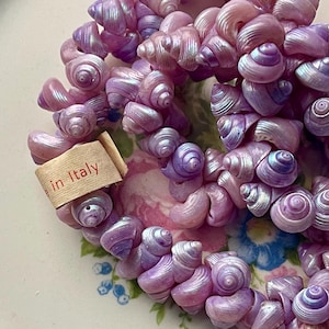 May include: A necklace composed of numerous small, iridescent spiral seashells in shades of light purple and lavender. A small, light-colored fabric tag is attached to the strand, featuring the printed text "made in Italy" in red ink.