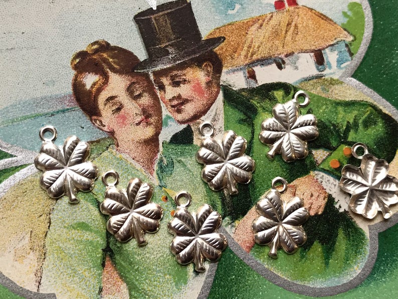 May include: Six silver four-leaf clover charms with a loop for hanging. The charms are on a green and white background with a vintage postcard image of a couple.