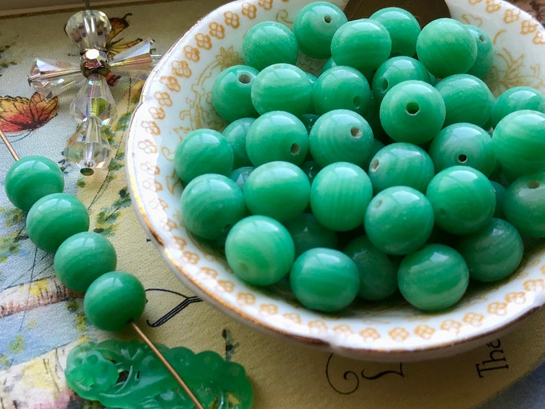 May include: A bowl filled with smooth, green jade beads. The beads are round and have a subtle swirl pattern.