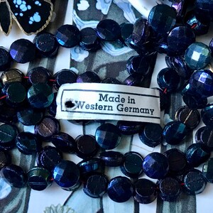 Puede incluir: Una hebra de cuentas de vidrio azul oscuro iridiscente con una etiqueta que dice "Made in Western Germany".