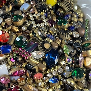 May include: A close-up of a collection of vintage jewellery components, including rhinestones, faux pearls, and various colourful gemstones. The components are in shades of red, green, blue, and yellow, with gold-toned metal settings. Ideal for jewellery making.