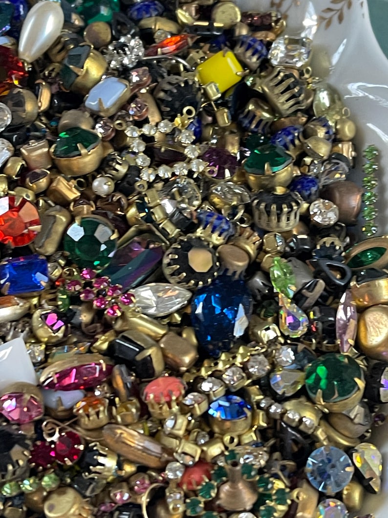 May include: A close-up of a collection of vintage jewellery components, including rhinestones, faux pearls, and various colourful gemstones. The components are in shades of red, green, blue, and yellow, with gold-toned metal settings. Ideal for jewellery making.