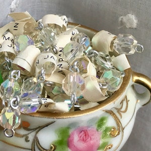 Puede incluir: Un peque&ntilde;o taz&oacute;n de porcelana blanca con ribetes dorados y un dise&ntilde;o floral, lleno de cuentas de vidrio transparentes iridiscentes y una cadena plateada.