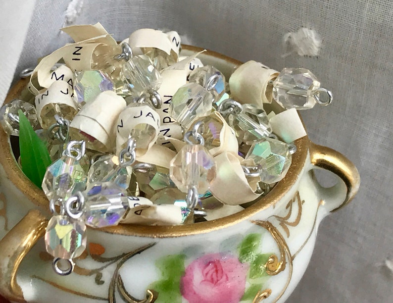 Puede incluir: Un peque&ntilde;o taz&oacute;n de porcelana blanca con ribetes dorados y un dise&ntilde;o floral, lleno de cuentas de vidrio transparentes iridiscentes y una cadena plateada.