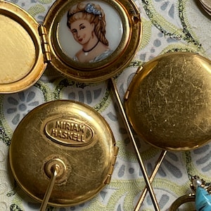 May include: Two gold-toned locket pins with a painted portrait inside one and the inscription "MIRIAM HASKELL" on the other.
