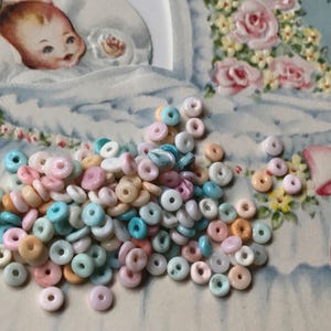 May include: A scattering of small, pastel-coloured, ring-shaped beads in shades of pink, blue, peach, and white. The beads are on a vintage baby-themed card with a baby illustration and floral accents.