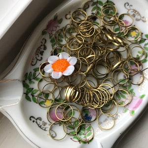Peut inclure: Un plat en c&eacute;ramique blanche avec un motif floral et une bordure dor&eacute;e. Le plat est rempli d'une grande quantit&eacute; de petits anneaux m&eacute;talliques dor&eacute;s. Une petite fleur blanche avec un centre orange repose sur les anneaux.