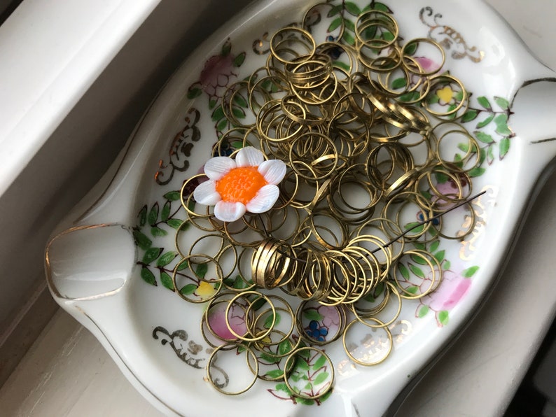 Peut inclure: Un plat en c&eacute;ramique blanche avec un motif floral et une bordure dor&eacute;e. Le plat est rempli d'une grande quantit&eacute; de petits anneaux m&eacute;talliques dor&eacute;s. Une petite fleur blanche avec un centre orange repose sur les anneaux.