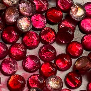 May include: A collection of red and iridescent glass gems. The gems vary in size and shape, with a mix of round and oval forms. The colors range from deep ruby red to lighter pink and shimmering gold. The reflective surfaces create a sparkling effect.