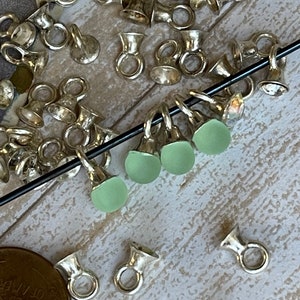 May include: A close-up of silver-toned metal findings with a small loop and a cup-shaped top. Four light green glass beads are strung on a black cord, threaded through the loops of the findings.