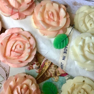 May include: A collection of carved rose-shaped beads in shades of pink and cream, with green leaf accents. Some beads have small holes for stringing. The image includes a ribbon that says "MADE IN JAPAN".