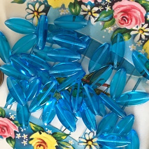 May include: A collection of approximately 30 turquoise blue glass beads. The beads are oval shaped and have a slightly iridescent finish.