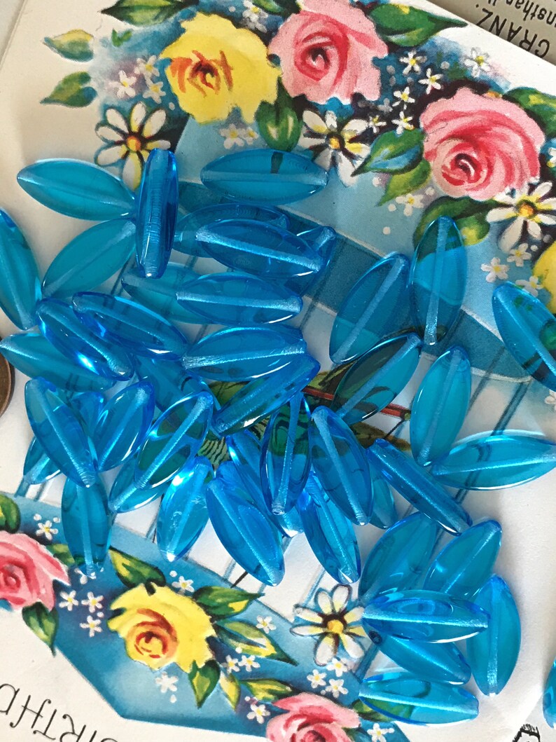May include: A collection of approximately 30 turquoise blue glass beads. The beads are oval shaped and have a slightly iridescent finish.