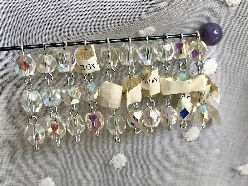 Puede incluir: Una hebra de cuentas de cristal iridiscente cuelga de un alambre plateado. Las cuentas son facetadas y tienen un brillo arco&iacute;ris. Un trozo de papel rasgado con el texto "MADE IN JAPAN" est&aacute; unido al alambre.