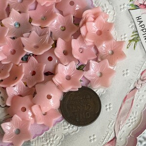 Peut inclure: Une collection de boutons en plastique rose en forme de fleur avec un seul trou au centre. Les boutons sont dispos&eacute;s sur une dentelle blanche avec une carte vintage en arri&egrave;re-plan.