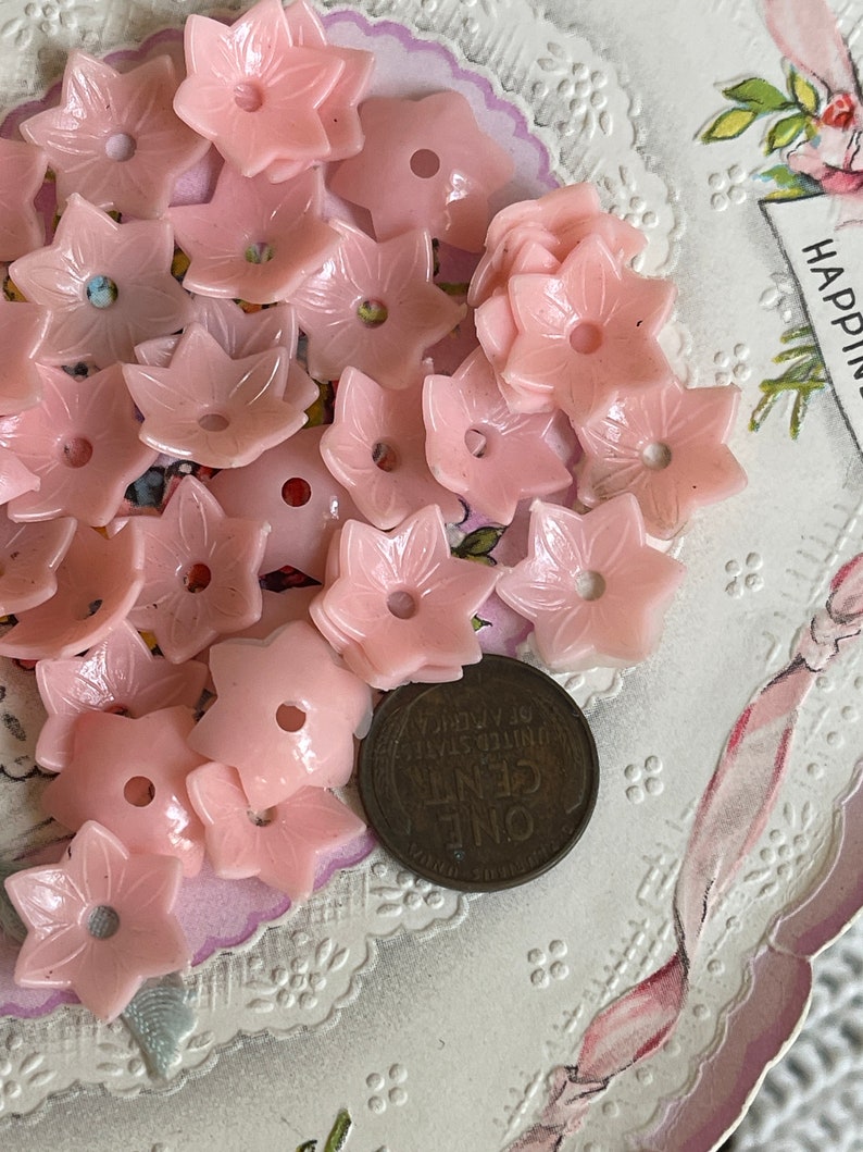 Peut inclure: Une collection de boutons en plastique rose en forme de fleur avec un seul trou au centre. Les boutons sont dispos&eacute;s sur une dentelle blanche avec une carte vintage en arri&egrave;re-plan.