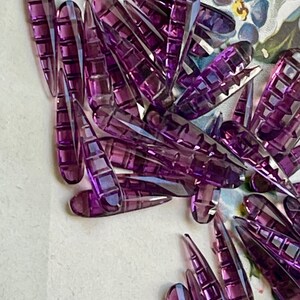 May include: A large group of purple glass teardrop-shaped beads with a faceted design. The beads are scattered on a white surface.