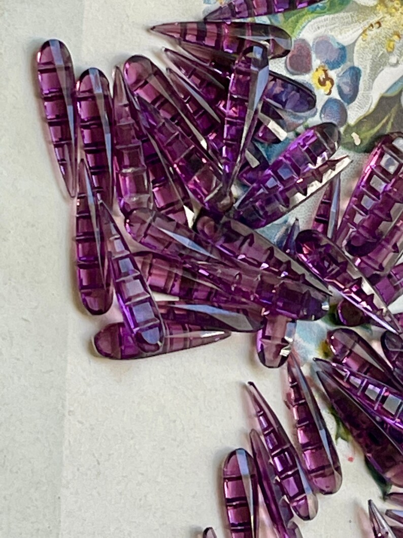 May include: A large group of purple glass teardrop-shaped beads with a faceted design. The beads are scattered on a white surface.
