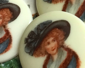 May include: Three round, white, ceramic buttons with a painted image of a woman wearing a black hat with a bow. The woman has red hair and is wearing a blue and white dress with a brown fur collar.